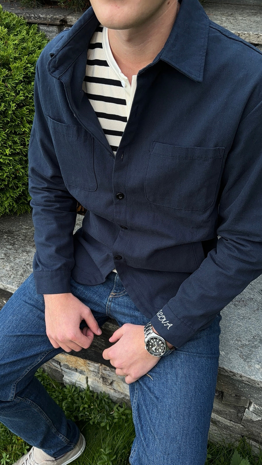 Sittende i Navy Overshirt, ASCOVA-brodering på mansjetten ved siden av en stålklokke