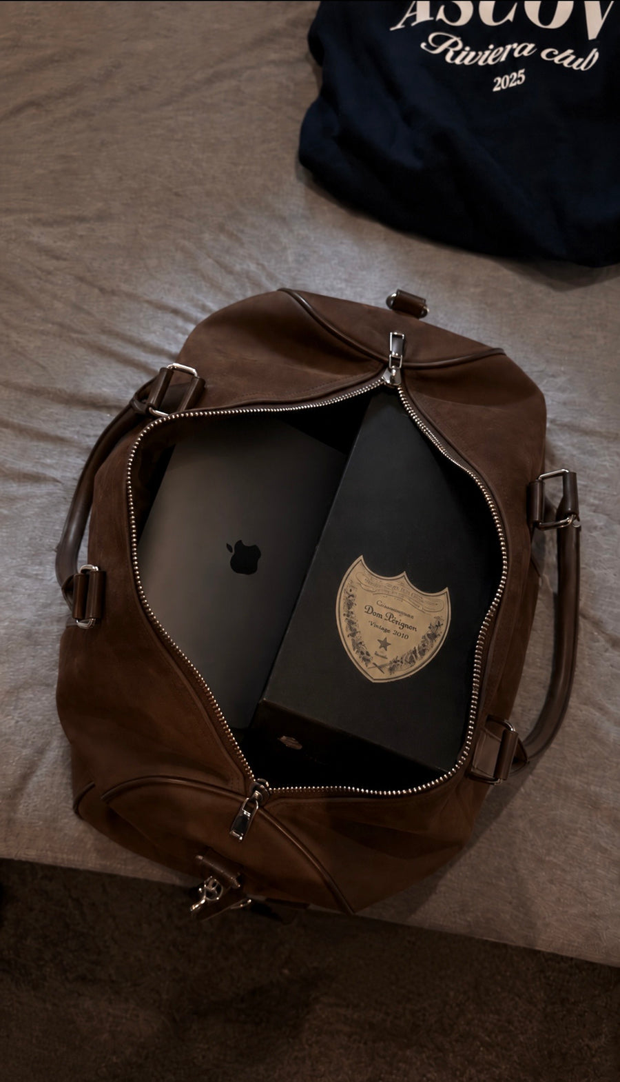Ascova Suede Duffel åpen på en seng, pakket med en laptop og en flaske champagne